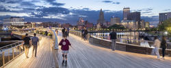 a person in a red hooded sweatshirt skates along a pedestrian bridge at night with the Providence skyline in the background and other people nearby