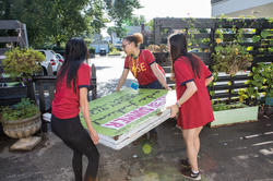 three students in red shirts carry a sign through a gate at a community garden
