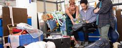 Three students huddle around a laptop while surrounded by suitcases and bins.