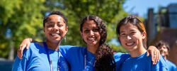 three smiling students in blue t-shirts huddle together outdoors in front of a tree with full green leaves