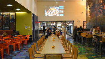 students eating at a long dining table, flanked by red furniture on the left and more tables on the right