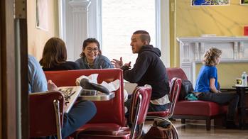 students sit together at a cafe table with other guests in the background in front of a yellow wall and sunlit window