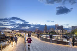 a person in a red hooded sweatshirt skates along a pedestrian bridge at night with the Providence skyline in the background and other people nearby