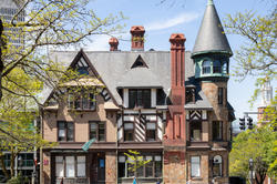 a light brown house with darker framed windows and a corner tower, surrounded by trees against a blue sky