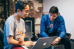 a student works on a laptop computer inside an office as a colleague looks on in the background