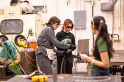 One person is holding a tool and talking to another over a workdesk.