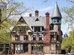 a light brown house with darker framed windows and a corner tower, surrounded by trees against a blue sky