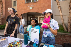 three students outdoors behind a table with a sign that reads “Health Education and Promotion”, all in front of a red-brick building
