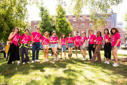 Group of RISD orientation leaders in pink shirts with a print that says "Hi" standing on a green with trees and a brick campus building behind them.