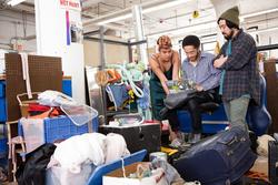 Three students huddle around a laptop while surrounded by suitcases and bins.