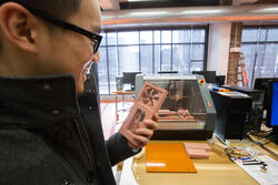 a student inside a well-lit workspace holds a brown, 3d-printed prototype object about the size of a smartphone