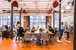 students with laptops sitting on both sides of a long table inside a workshop, surrounded by equipment