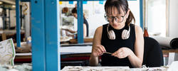 a student wearing glasses and a pair of headphones around their neck concentrates on work while seated at a studio table