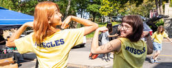 two students flex outdoors on a sunny day while showing the "Muscles" logo on the back of their t-shirts