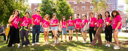 Group of RISD orientation leaders in pink shirts with a print that says "Hi" standing on a green with trees and a brick campus building behind them.