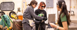 One person is holding a tool and talking to another over a workdesk.