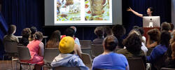 A group of RISD graduate students attend an info session where the speaker is presenting information to the group on a projector.