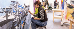 A RISD student is sitting on a stool and working on a large visual artwork in one of our studio spaces.
