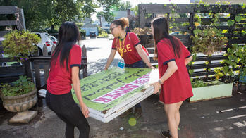 three students in red shirts carry a sign through a gate at a community garden