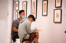 Two students sit looking at a R I S D gallery wall while sketching and taking notes
