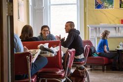 students sit together at a cafe table with other guests in the background in front of a yellow wall and sunlit window