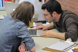 Two people lean across the table to look at something on a computer.