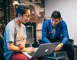 a student works on a laptop computer inside an office as a colleague looks on in the background