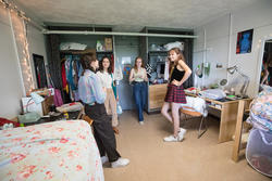 four students stand inside a college dorm room, in front of two closets with many clothing items visibly hanging