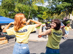 two students flex outdoors on a sunny day while showing the "Muscles" logo on the back of their t-shirts