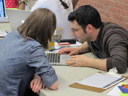 Two people lean across the table to look at something on a computer.