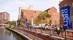 A crowd of people walk alongside the Providence River where RISD banners are waving.